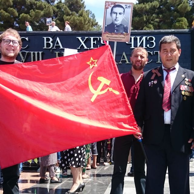 Ein Mann mit vielen Orden posiert stolz neben einer Sowjetflagge.