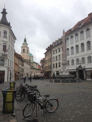 Eine leere Straße in Ljubljana mit Kirchentürmen im Hintergrund und abgestellten Fahrrädern im Vordergrund.