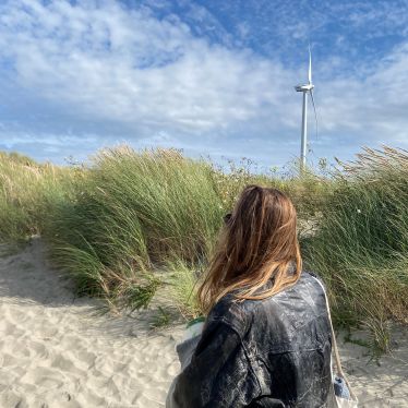 Man sieht mich während ich an den Dünen am Strand entlang laufe. In Hintergrund ist ein Windrad und der Himmel ist blau mit ein paar Wolken.
