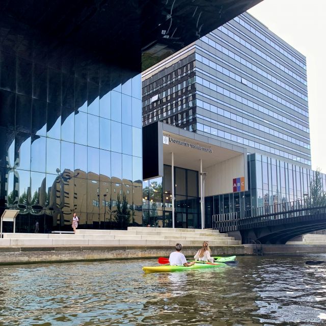 Man sieht ein orangenes Kanu auf einem Kanal. Der Kanal geht durch den Campus der Universität von Amsterdam. Links vom Kanal kann man große, gläserne Unigebäude erkennen.