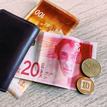 Im Zentrum des Bildes ist eine rosafarbene Banknote im Wert von 20 Schekel zu sehen. Rechts am Rand sind zwei Münzen: 1/2 Schekel und 10 Agorot sowie eine 100 Schekel Banknote im Hintergrund.