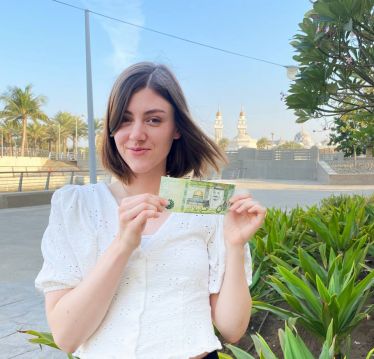Eine junge Frau hält einen 50 Riyal Gelschein hoch un dlächelt. im hingtegrund ist eine Moschee und Pflanzen zu sehen