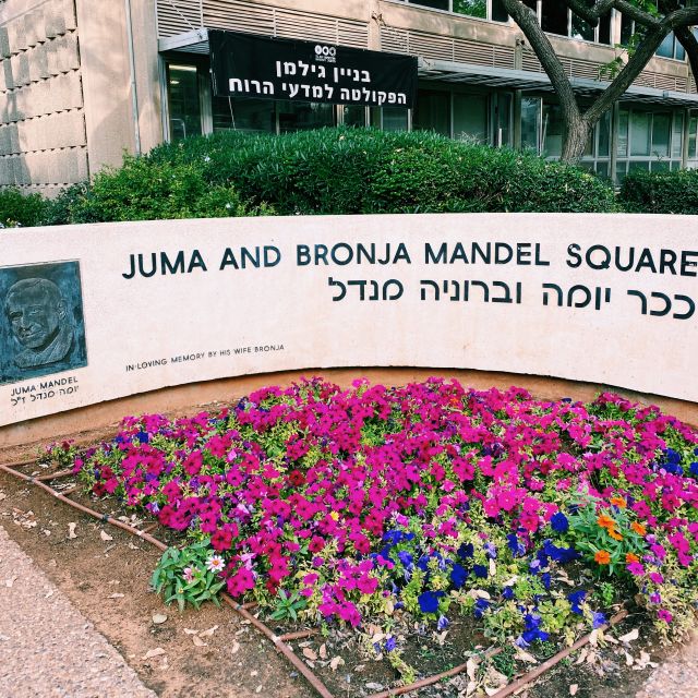 Im Vordergrund sind pinke Blumen auf einem Beet und mittig die Aufschrift "Juma and Bronja Mandel Square" in lateinischem und hebräischem Alphabet.