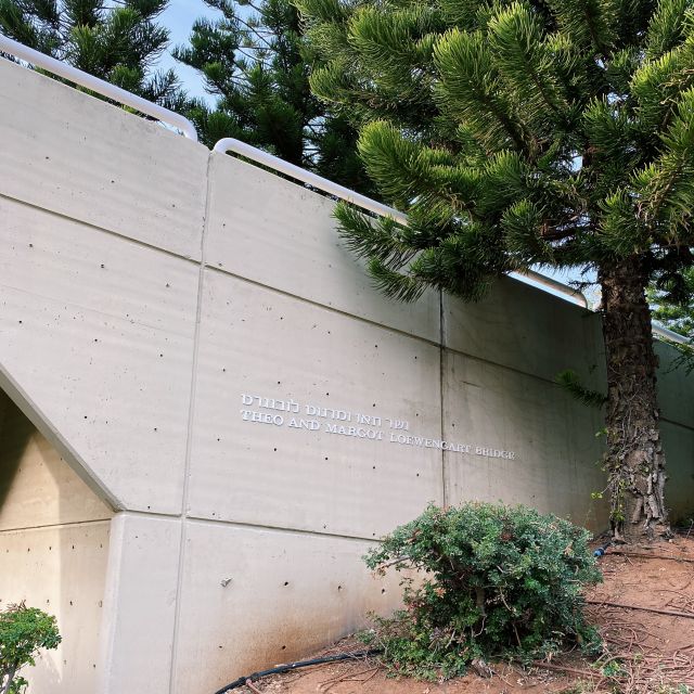 Zu sehen ist eine Wand von einer Brücke mit der Aufschrift "Theo and Margot Loewengart Bridge". Rechts im Bild ist ein grüner Baum.