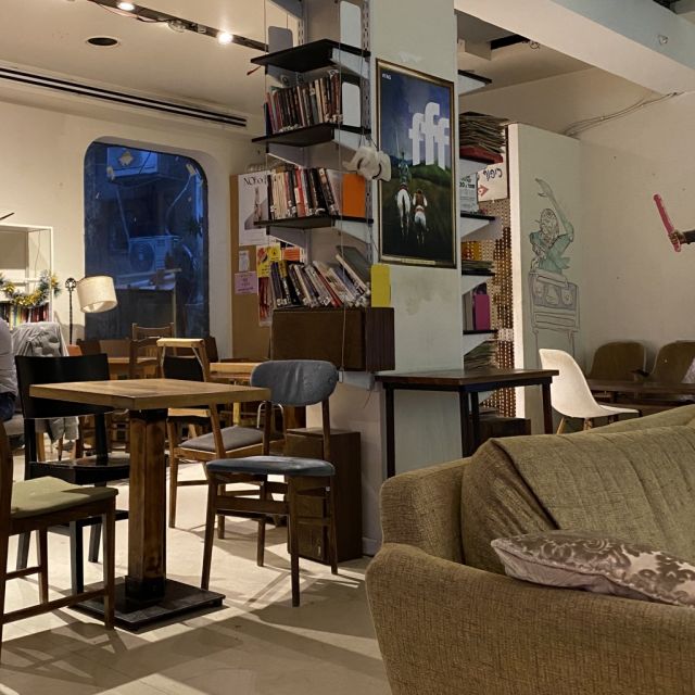 Auf dem Foto ist die Inneneinrichtung von Kiosko, einem Cafe in Tel Aviv zu sehen. Im Vordergrund rechts auf dem Bild ist ein Ausschnitt eines Sofas, mittig ein Bücherregal und links ein Tisch mit Stühlen.