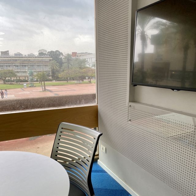 Zu sehen ist ein Ausschnitt eines runden Tisches mit einem Stuhl, rechts ein großer Bildschirm und im Hintergrund der Uni-Campus.