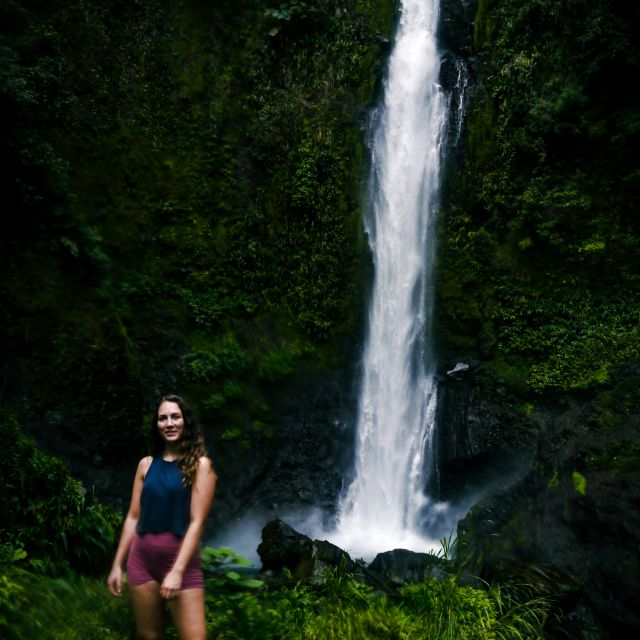 Carla steht in sportlicher Kleidung vor einem hohen Wasserfall in grün bewachsener Umgebung.