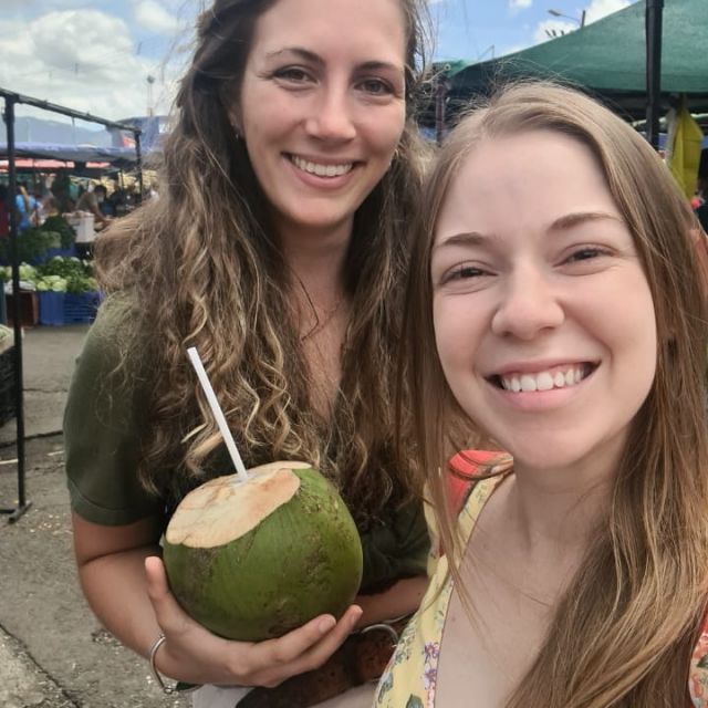 Carla und Sofia machen ein Selfie auf dem Markt, Carla hat eine grüne Kokosnuss in der Hand.