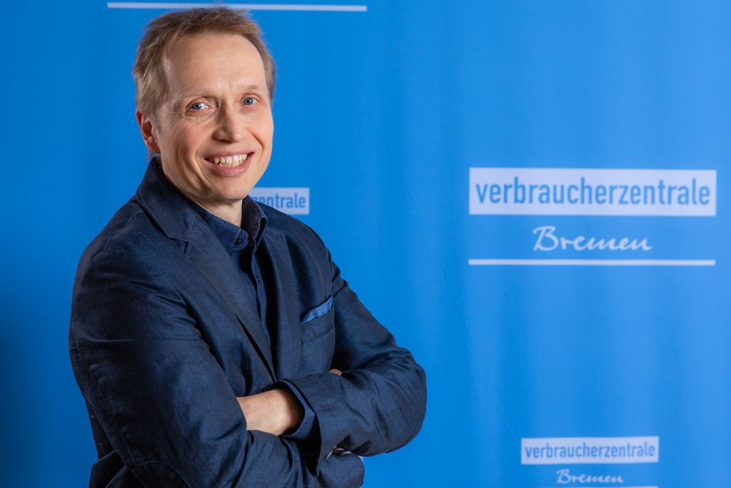 Portraitfoto von Finanzexperte Thomas Mai vor einer blauen Stellwand mit Aufdruck "Verbraucherzentrale".