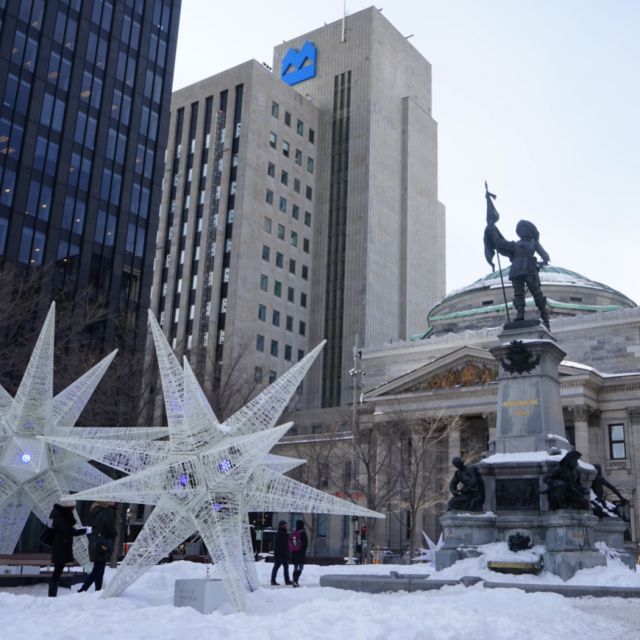 Eine Statue im Vordergrund. Im Hintergrund steht ein Theater. Der Platz ist in Schnee gehüllt. Vor der Statue stehen 2 große weiße Sterne