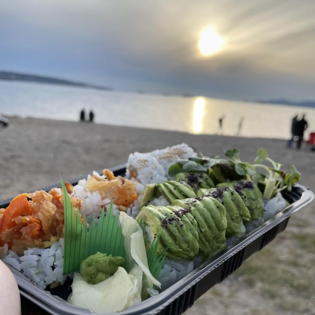 Eine Hand hält eine Schale mit verschiedenem Sushi in der Hand. Im Hintergrund geht die Sonne unter. Die Person sitzt am Strand.