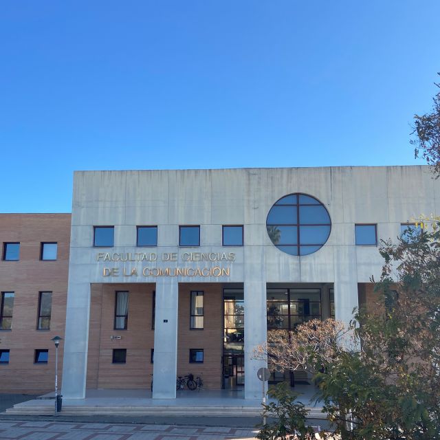 Das Gebäude der Facultad de Ciencias de la Comunicaión und Facultad de Turismo in frontaler Ansicht. Auf der linken Seite befindet sich der Schriftzug mit dem Namen der Fakultät. Das Gebäude steht unter strahlend blauem Himmel