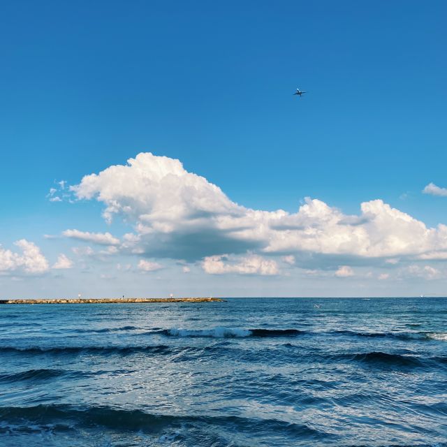 Auf dem Foto ist im unteren Bilddrittel das Meer zu sehen.Links ist ein schmaler Streifen der Küste und oberhalb der Himmel mit einer großen Wolke und einem Flugzeug in der Ferne zu sehen.