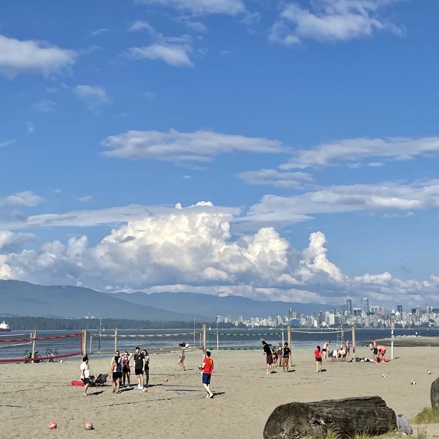 Sandstrand mit Menschen, die Beachvolleyball spielen. Dahinter die Innenstadt von Vancouver mit vielen Hochhäusern.
