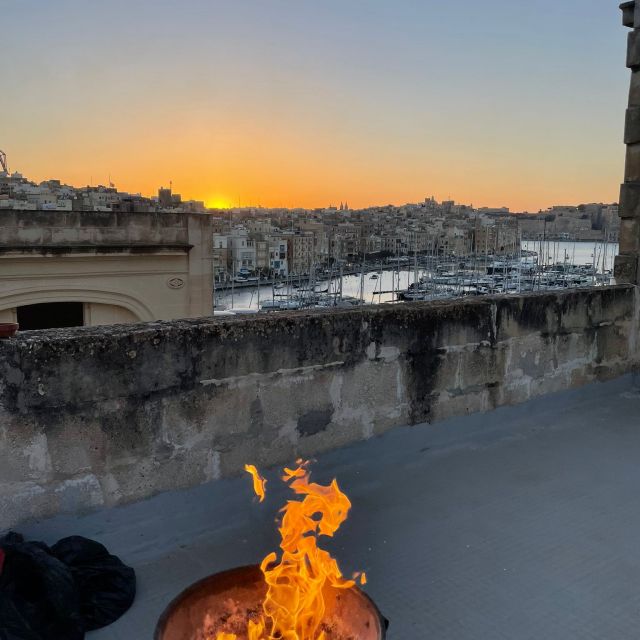 Ein brennendes Lagerfeuer auf einer Dachterrasse, im Hintergrund geht die Sonne vor Häuserfassaden und Booten unter.