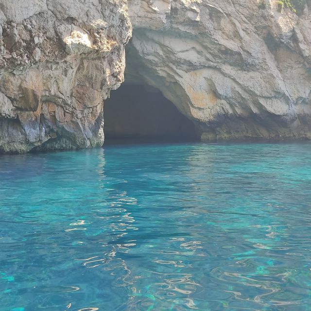 Schimmernd blaues Wasser vor sandsteinfarbenen Felsen mit Eingang zu einer Höhle