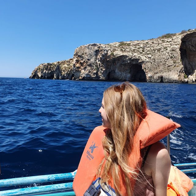 Ein blondes Mädchen mit oranger Schwimmweste sitzt in einem Boot und blickt auf das dunkelblaue Meer. Im Hintergrund sind Fels- und Höhlenformationen zu erkennen.