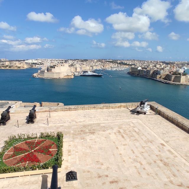 Ein Blick von oben auf einen großen Platz, auf dem sich ein wie das maltesische Wappen angeordnetes Blumenbeet und mehrere Kanonen befinden. Hinter dem Platz liegt das Meer und hinter dem Wasser sind weitere Städte und Häfen zu erkennen.