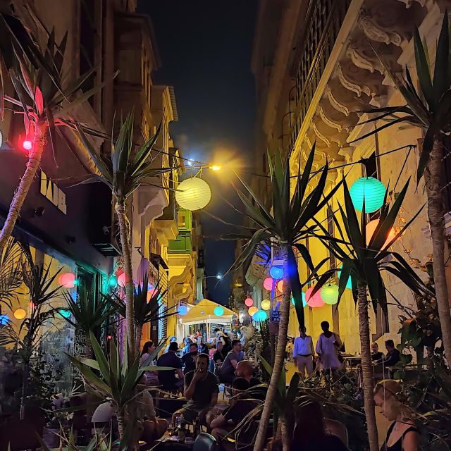 Eine Bar bei Nacht in einer schmalen Gasse mit vielen Menschen. Im Vordergrund stehen Palmen und es hängen überall bunte Lichterketten.