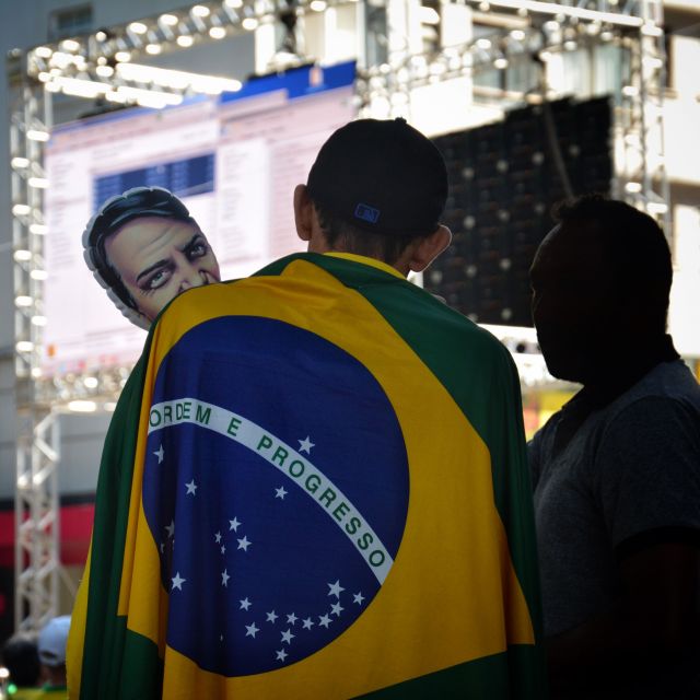 Ein Bolsonaroanhänger steht auf einer Demo, eingehüllt in eine Brasilienflagge und mit einer Bolsonaropuppe