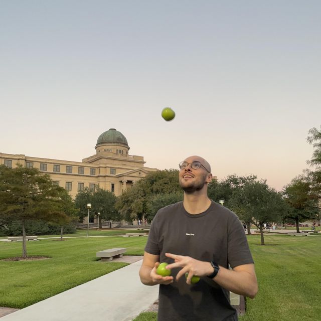 Ich stehe gekleidet in einem grauen T-Shirt und langer schwarzer Hose vor einem der schönsten Gebäude der Universität. Ich jongliere auf dem Bild einen grünen Apfel um mein bildliches Beispiel aus dem Text zu illustrieren.