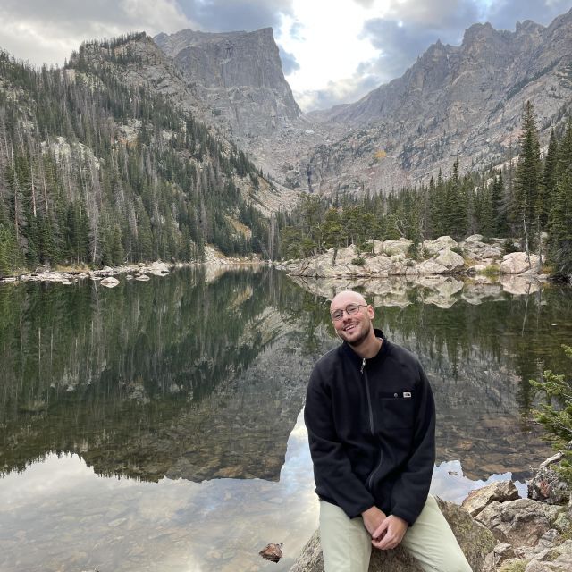 Ich sitze auf einem großen Stein. Ich habe eine Grüne Hose und einen schwarzen Vlies Pullover an. Im Hintergrund sind die Rocky Mountains zusehen und ein großer grüner See.