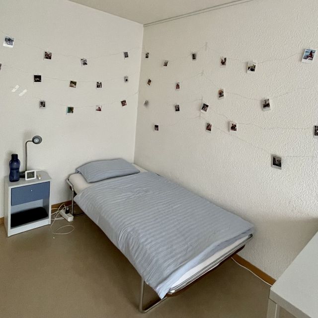 Ausschnitt aus Tobias' Zimmer.Ein Bett, ein Nachtkasten und an der Wand viele Polaroids mit durchsichtigen Wäscheklammern an zwei Lichterketten hängend.