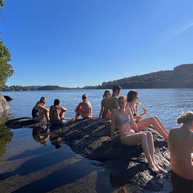 Mehrere Leute sitzen in Badekleidung auf Steinen, im Hintergrund ein Fjord.