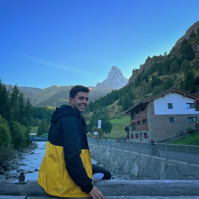 Tobias sitzt auf einer Brückenhalterung. Hinter ihm sieht man einen kleinen Fluss und das Matterhorn.