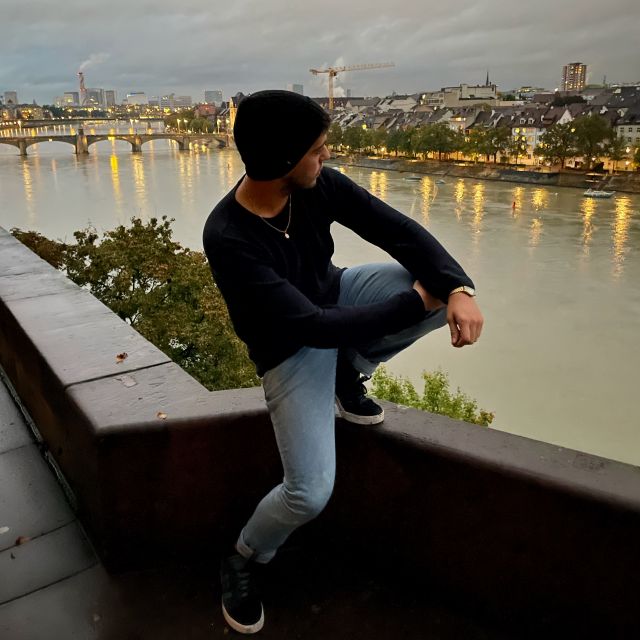 Tobias steht in Basel an einer Aussichtsplattform am Abend. Er schaut auf die gegenüberliegende Seite des Rheins. Die Lichter der Stadt leuchten bereits auf. Mit dem bewölkten Himmel und der Rauchwolken der Fabriken kommt eine eher düstere Stimmung auf.