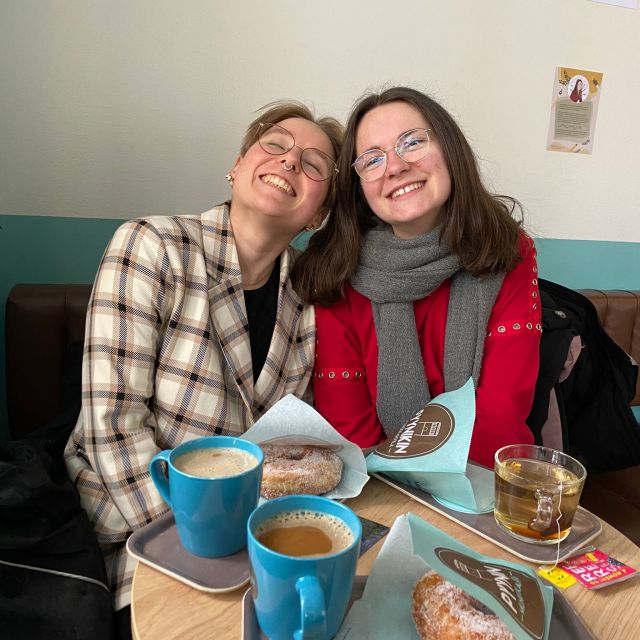 Zwei junge Frauen sitzen im Café, auf dem Tisch vor ihnen stehen Kaffee, Tee und Gebäck, beide lächeln in die Kamera