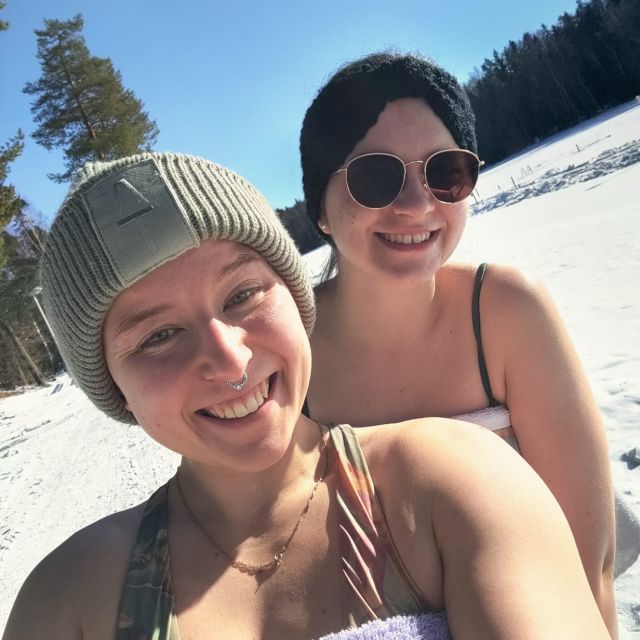 Zwei junge Frauen in Bikinis und Handtüchern lächeln in die Kamera, die Sonne scheint und im Hintergrund liegt Schnee