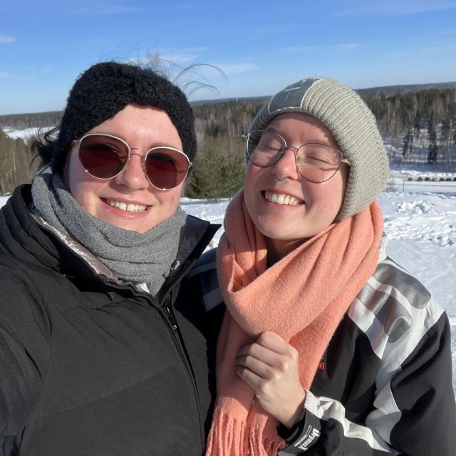 Zwei junge Frauen in Winterkleidung lächeln in die Kamera, die Sonne scheint und es liegt Schnee