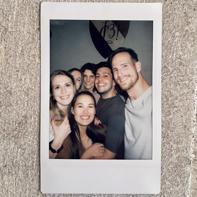 Ein Polaroid mit 6 Menschen drauf. Einer davon Tobias, umzingelt von vier Frauen und einem Mann. Alle lächeln glücklich in die Kamera.