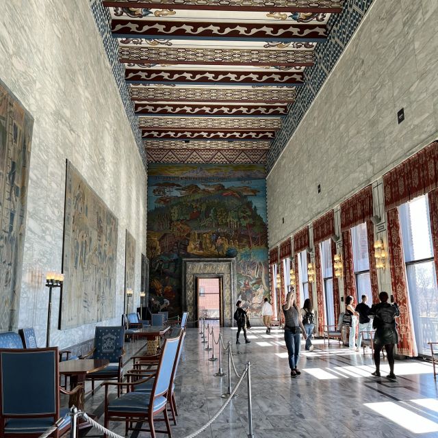 Langer Gang mit hoher Decke und Fensterfront, an den Wänden hängen Kunstwerke und die Decke ist mit einem bunten Muster verziert