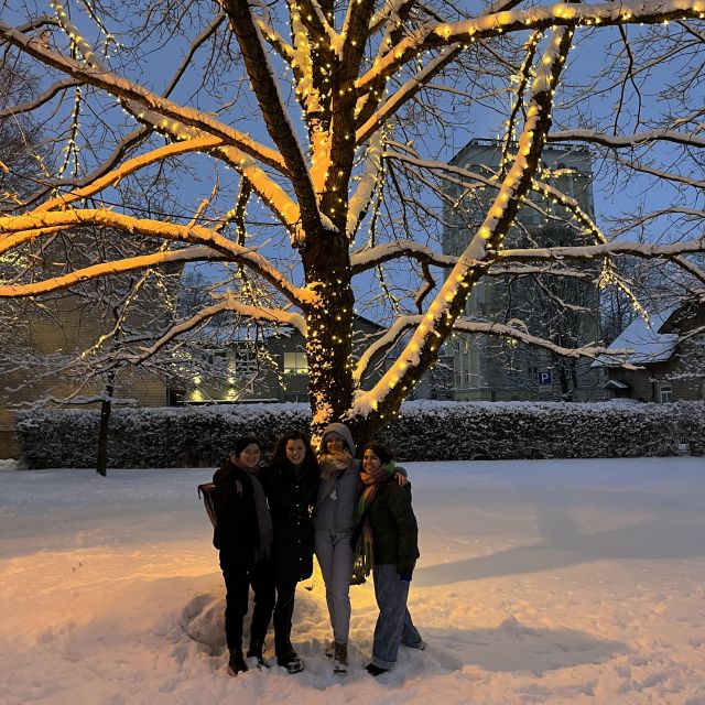 Eine Gruppe von vier jungen Frauen steht vor einem beleuchteten Baum im Schnee, der Himmel ist fast dunkel.
