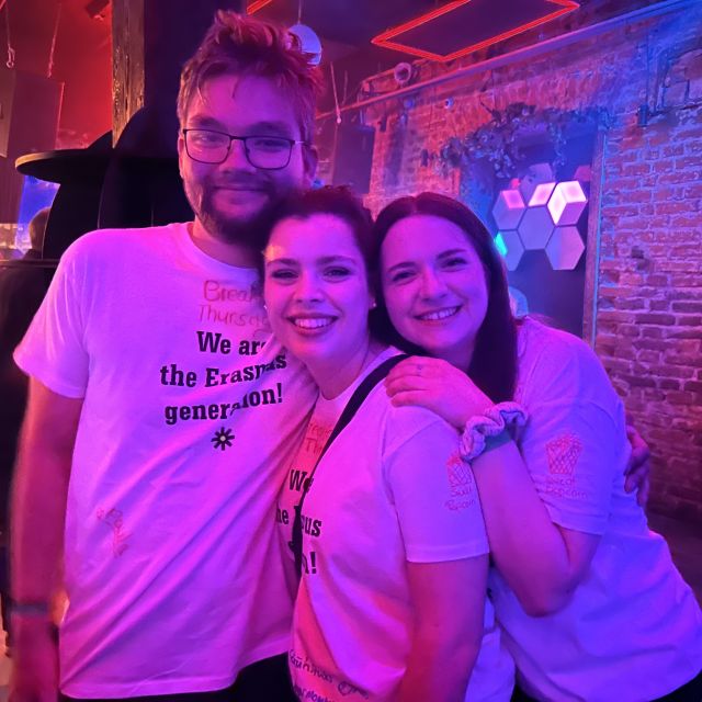 Zwei junge Frauen und ein junger Mann stehen gemeinsam in einem Club. Sie sind in violettes Licht getaucht und tragen alle ein weißes T-Shirt und lächeln in die Kamera.
