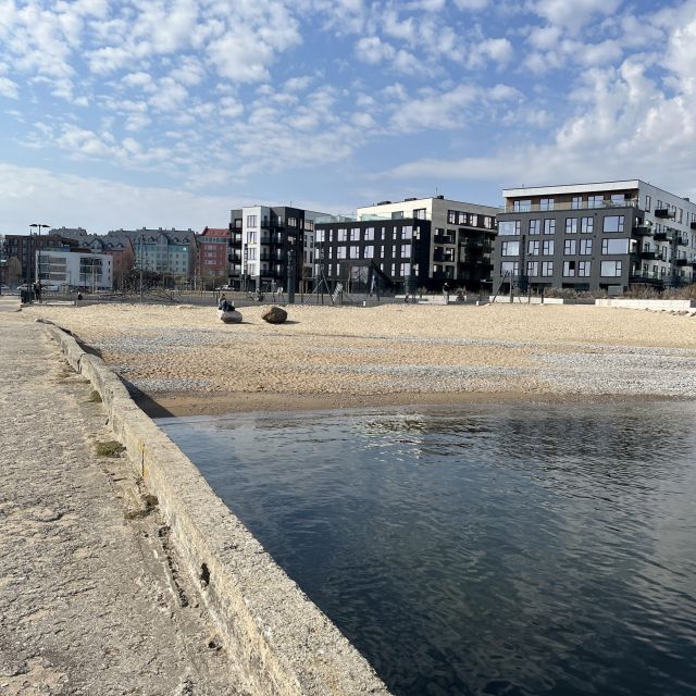 Promenade mit modernen Appartment-Gebäuden, davor ein kleiner Sandstrand und etwas Meer, darüber blauer Himmel mit Wölkchen