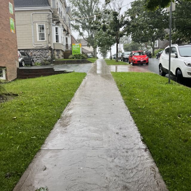 Blick auf einen Gehweg, der vertikal etwas bergauf verläuft, links ist eine Häuserfront. Wasser läuft den Weg hinab, es regnet in Strömen.