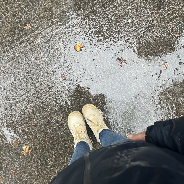 Vogelperspektive auf Schuhe im Regen. Man sieht eine Pfütze auf dem Boden, Jeans und Jacke sind durchnässt.