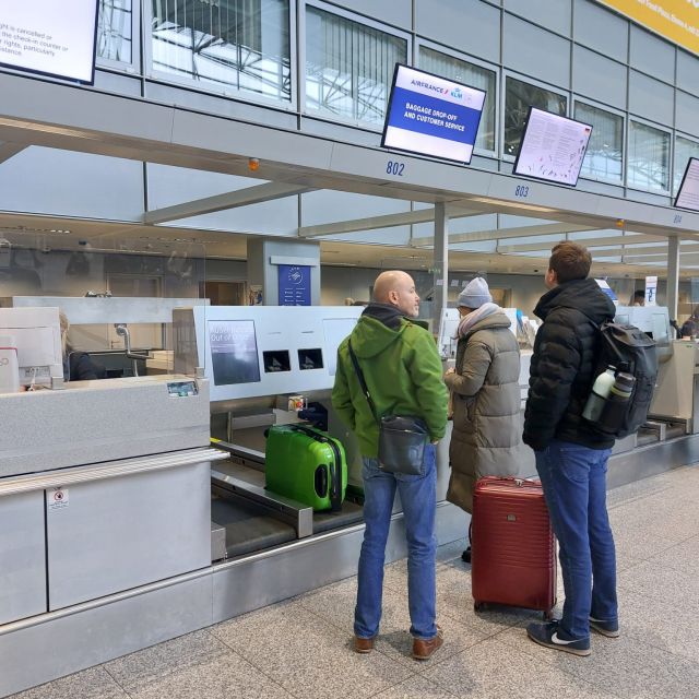 Bei der Gepäckabgabe am Flughafen, drei Menschen am Schalter stehend. Ein Koffer auf dem Band, ein weiterer auf dem Boden stehend.