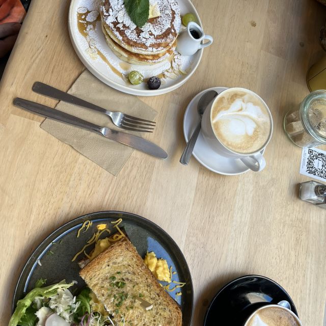 Bild von oben auf zwei Kaffees und zwei Teller mit Essen auf einem Holztisch. Auf einem Teller sind Pancakes mit Puderzucker, auf dem anderen ein Grilled Cheese Toast mit Salat.