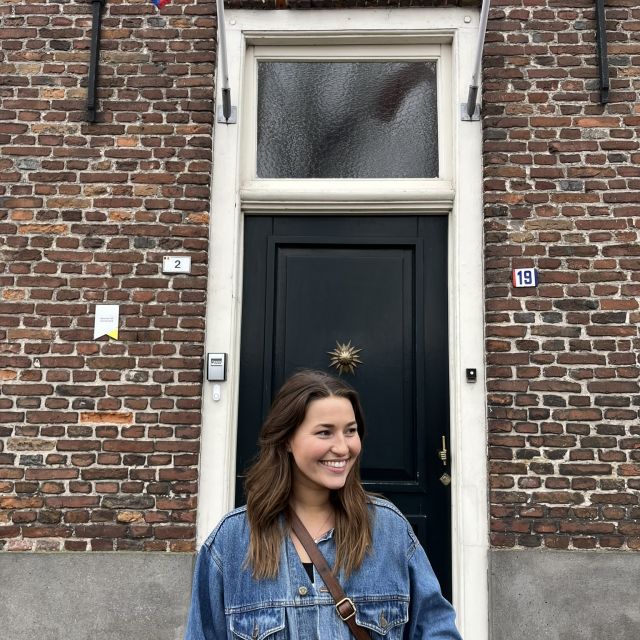 Im Vordergrund steht eine Frau in Jeansjacke, der Blick zur Seite. Sie lächelt und steht vor einer Tür eines Backsteinhauses.