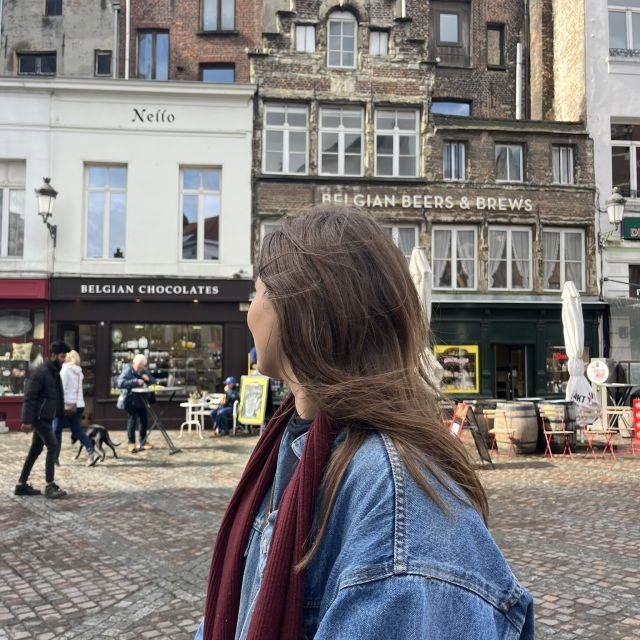 Eine Frau steht im Vordergrund; sie trägt eine Jeansjacke, einen roten Schal und lange offene Haare. Blick nach hinten auf eine Häuserfassade aus Backstein. Ein Haus ist ein Schokoladen-Laden, im anderen wird belgisches Bier verkauft.