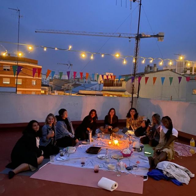 Auf einer Dachterrasse mit Lichterkette sitzen eine Reihe an Menschen im Kreis und picknicken